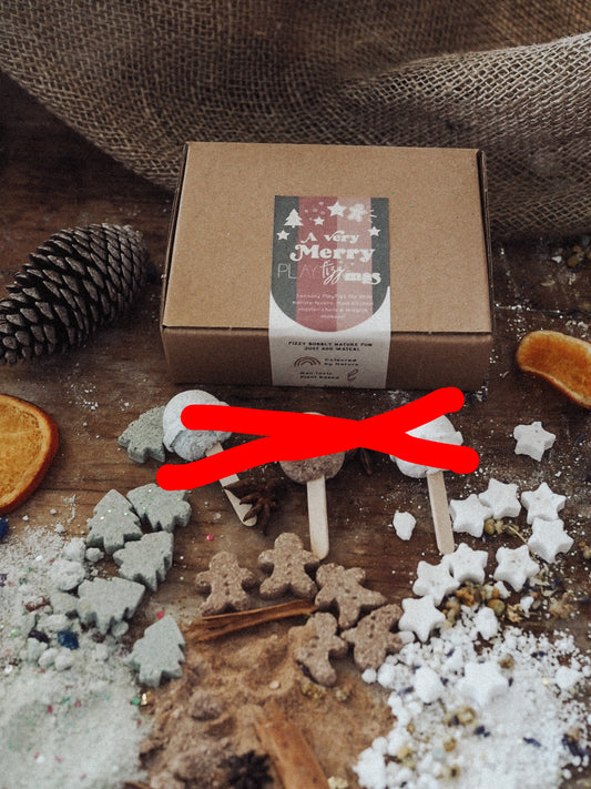 The image shows a collection of natural ingredients for PlayFizz activities, including dried flowers, spices, and crystals, with a cardboard box labeled 'Merry Fizzmas' in the background.